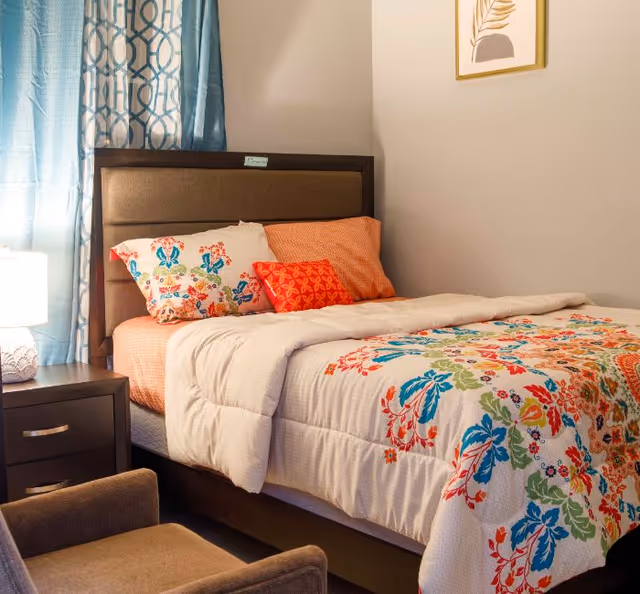 A cozy bedroom with a bed featuring a colorful floral-patterned comforter and multiple pillows in orange and floral designs. Next to the bed is a dark wooden nightstand with a white lamp. Blue patterned curtains hang by the window, and a framed artwork is on the beige wall. A brown upholstered chair is partially visible in the foreground.