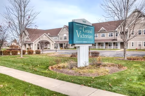 Exterior view of the Grand Victorian senior living facility showing a large building with multiple windows and a peaked roof. In the foreground, there is a green sign with white text that reads 'The Grand Victorian' along with the address number 1440. The surrounding area includes a sidewalk, grass, some bushes, and leafless trees.