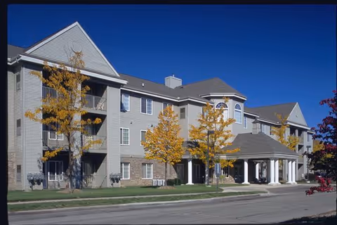 Exterior view of a multi-story senior living facility building with beige siding and a covered entrance supported by white columns. There are several trees with yellow autumn leaves in front of the building under a clear blue sky.