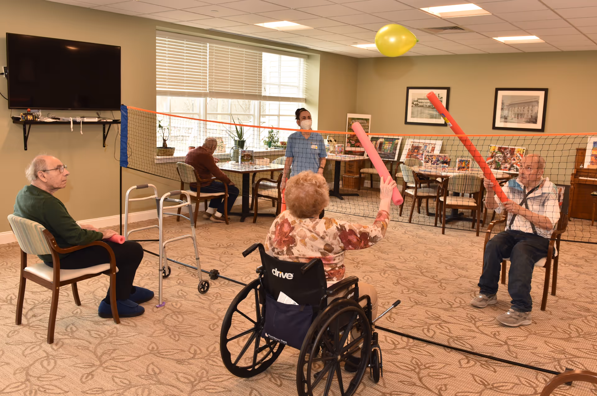 A group of elderly individuals playing seated volleyball indoors using pool noodles to hit a balloon over a net. One person is in a wheelchair, and a staff member wearing a mask is standing nearby. The room has beige walls, a large window, a television mounted on the wall, and several tables and chairs in the background.