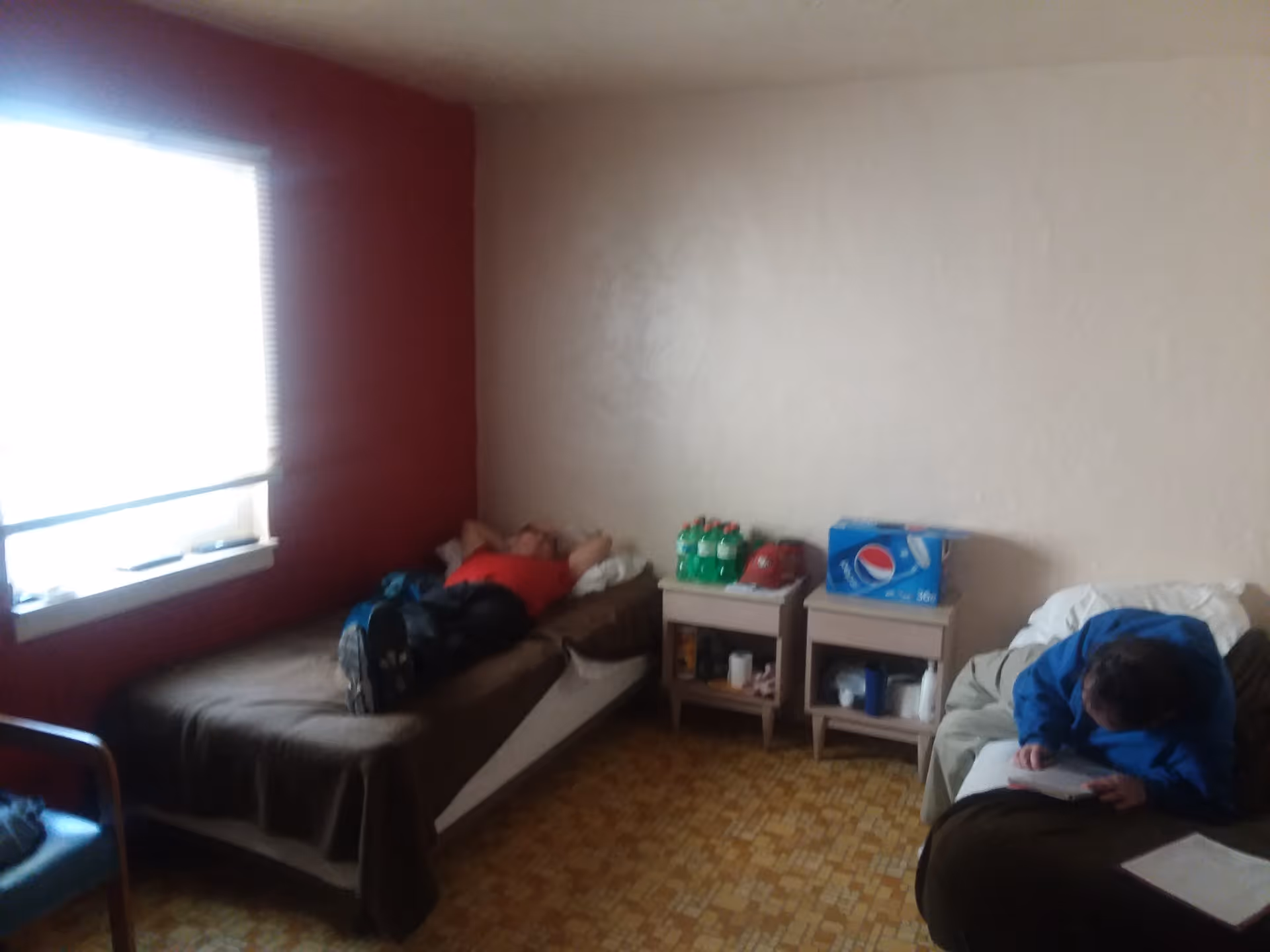 A small room with two single beds, one on the left side with a person lying down wearing a red shirt and dark pants, and another on the right side with a person sitting and writing on a piece of paper. Between the beds are two small nightstands holding soda bottles, a box of Pepsi, and other miscellaneous items. The room has a window with blinds on the left wall and a patterned floor.