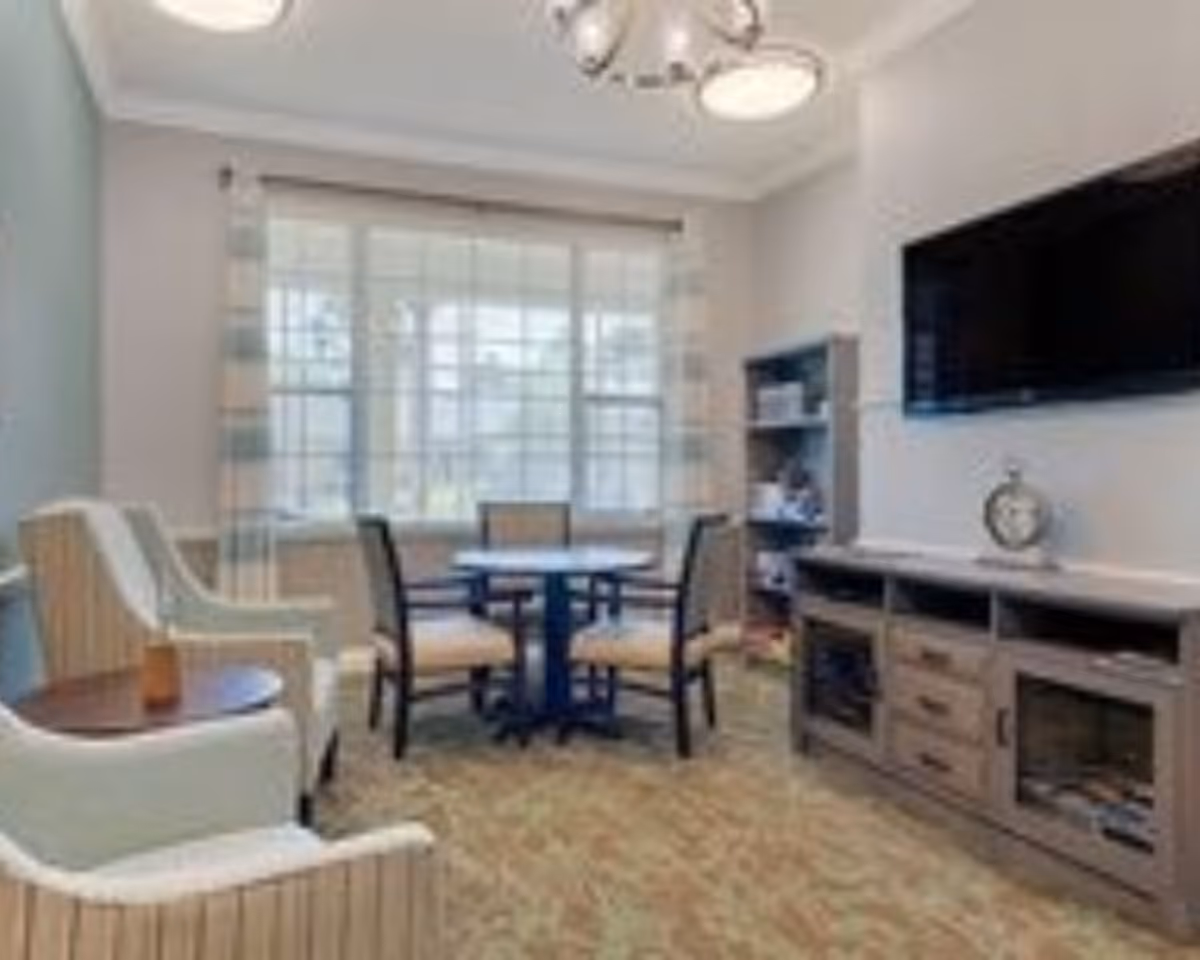 A cozy living room area with a round table and four chairs near a large window with sheer curtains. There are two armchairs with a small round side table between them on the left. On the right side, there is a wooden entertainment unit with a flat-screen TV mounted above it and decorative items on the shelves.