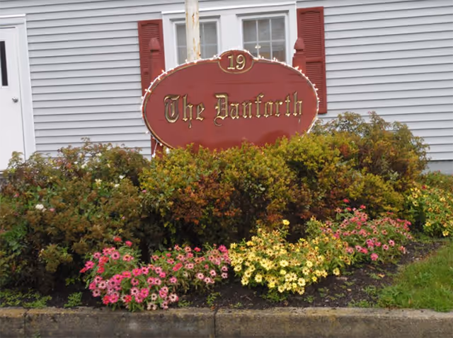 A red oval sign with gold lettering that reads 'The Danforth' and the number 19 above it, surrounded by green bushes and colorful flowers in front of a gray building with white trim and red shutters.