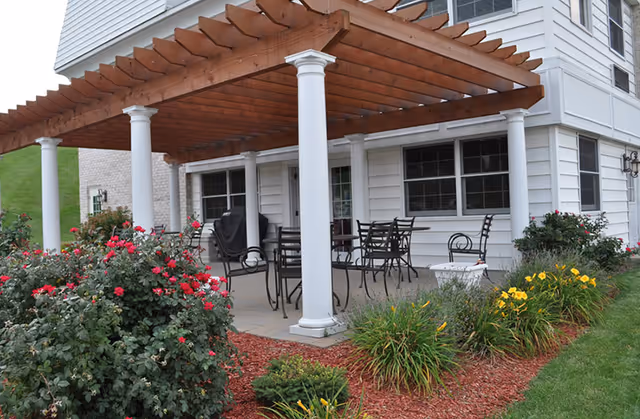 Outdoor patio area with a wooden pergola supported by white columns, metal chairs and tables arranged underneath, surrounded by flower beds with red and yellow flowers and green shrubs, adjacent to a white building with windows.