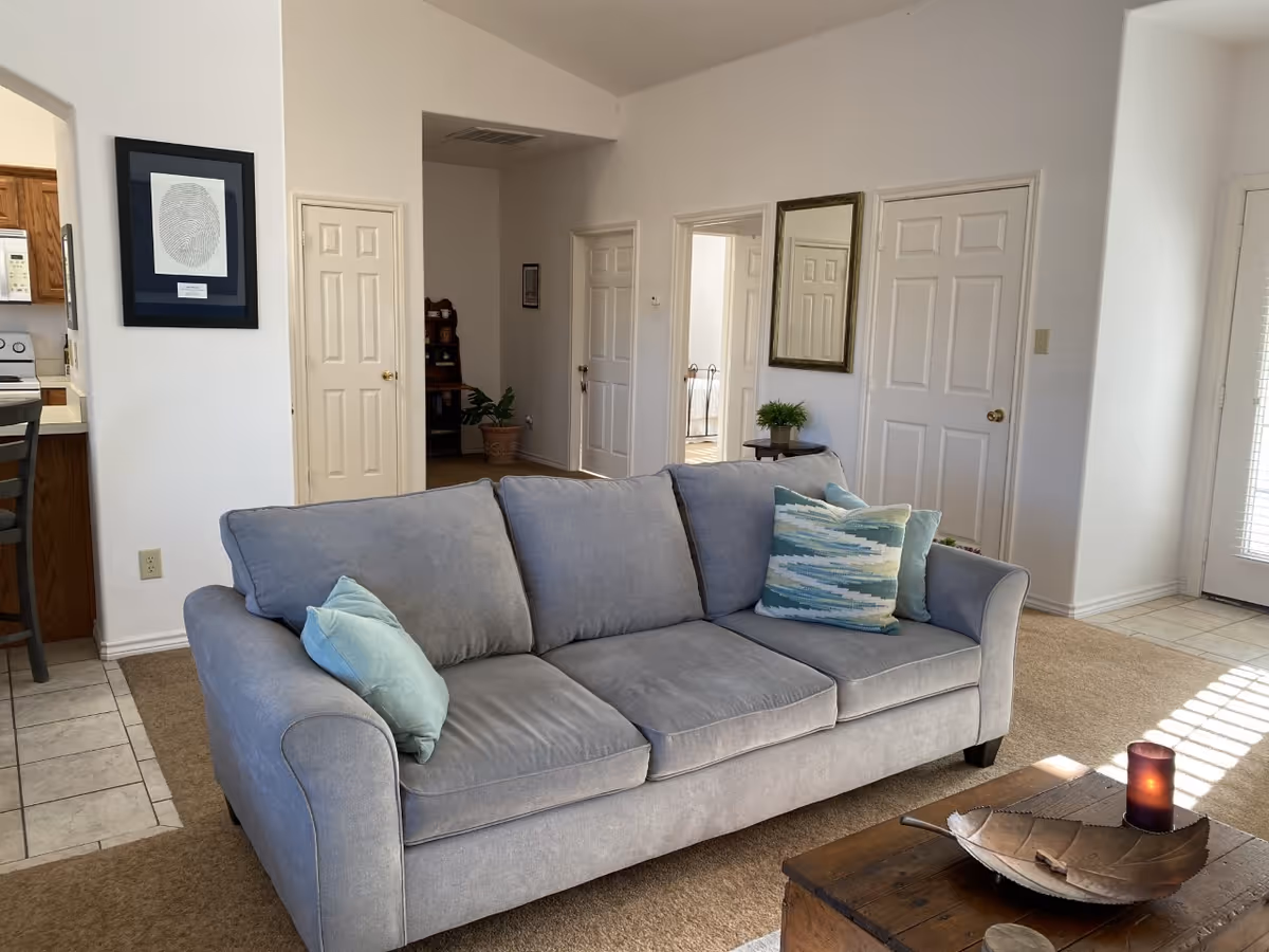 Sunny living room with a gray three-seat sofa, patterned throw pillows, and a wooden coffee table in front of multiple interior doors.