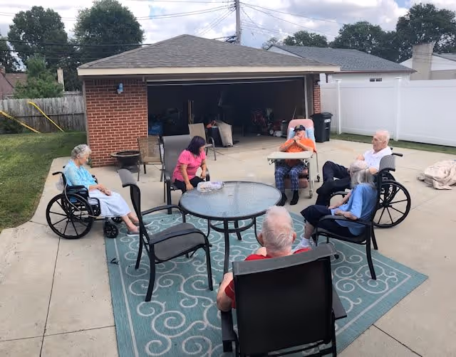 A group of elderly people sitting outside on a concrete patio in front of a garage. They are seated in wheelchairs and regular chairs around a glass-top round table on a blue patterned outdoor rug. The background shows a wooden fence, a white vinyl fence, and some trees under a partly cloudy sky.