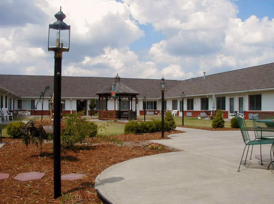 Outdoor courtyard area of a senior living facility with a paved walkway, black lamp posts, green metal chairs and tables, landscaped garden beds, and a wooden gazebo in the center. The building surrounds the courtyard with white siding and a brown roof under a partly cloudy sky.