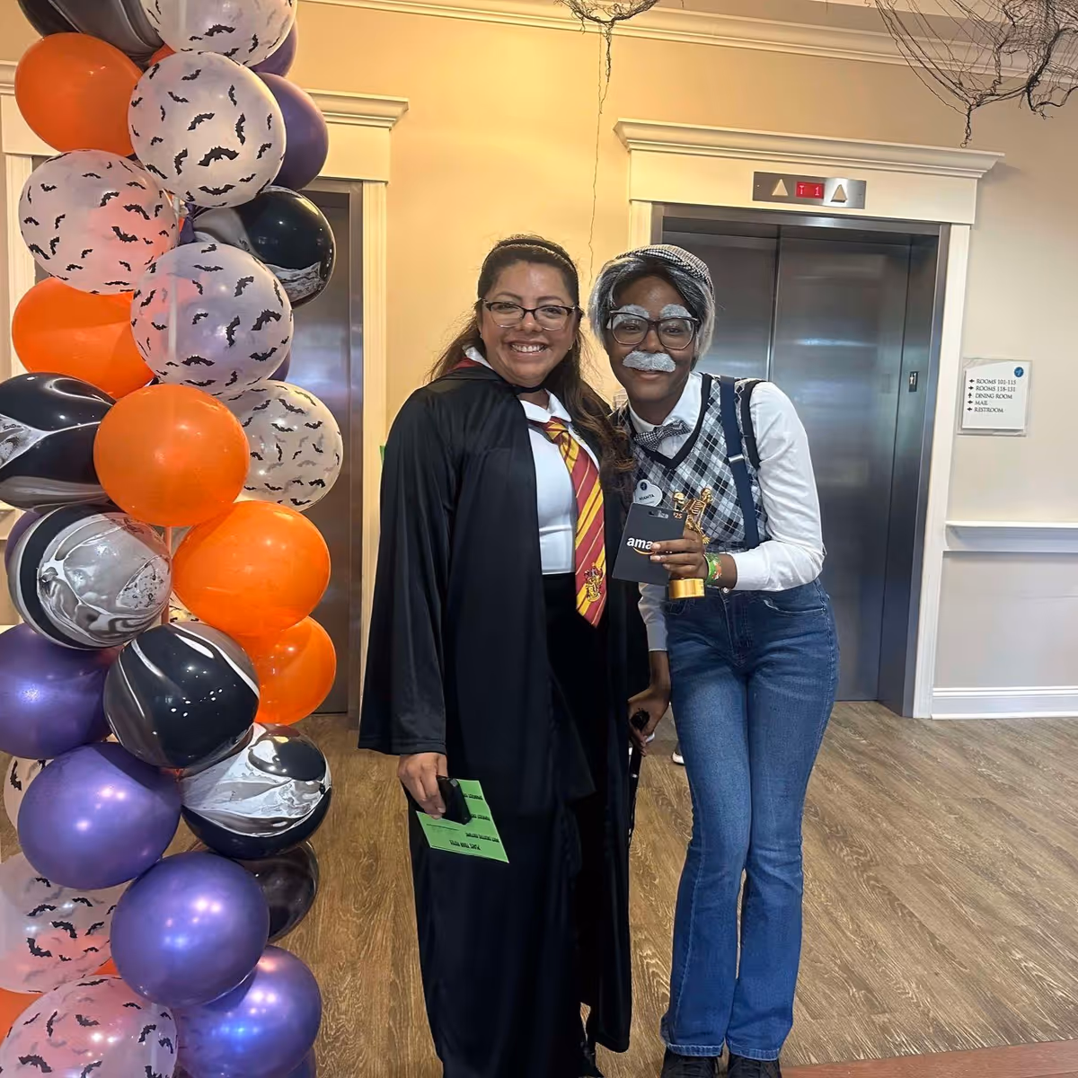 Two women standing indoors in front of elevators, one dressed in a Harry Potter costume with a black robe and Gryffindor tie, and the other dressed as an elderly person with gray hair, glasses, and a fake mustache, holding a small trophy and an Amazon gift card. There is a colorful balloon arch with orange, purple, black, and white balloons decorated with bat patterns on the left side.