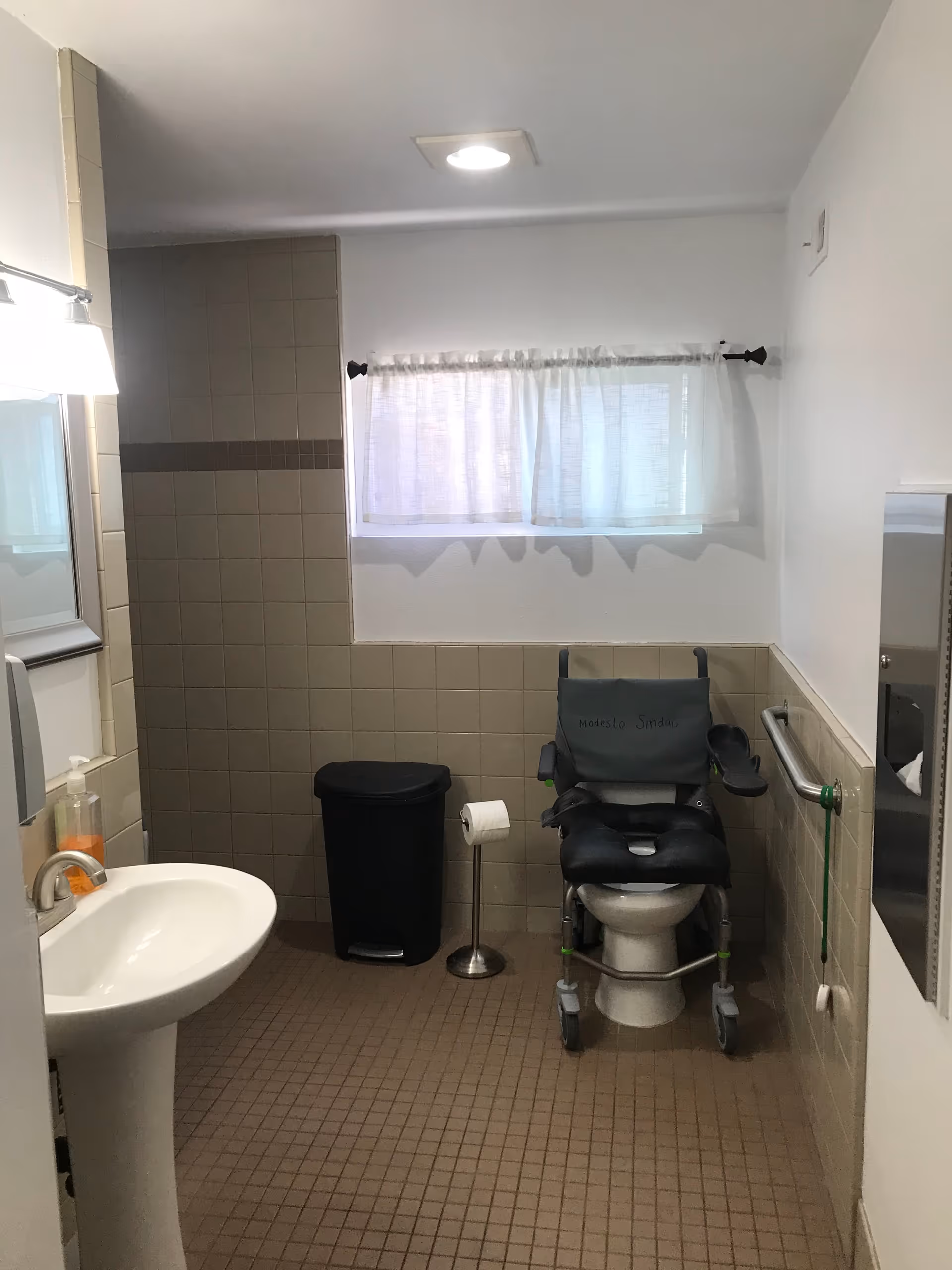 Accessible bathroom with a pedestal sink, a toilet fitted with a wheeled commode chair, grab bars, trash can, and a small curtained window.