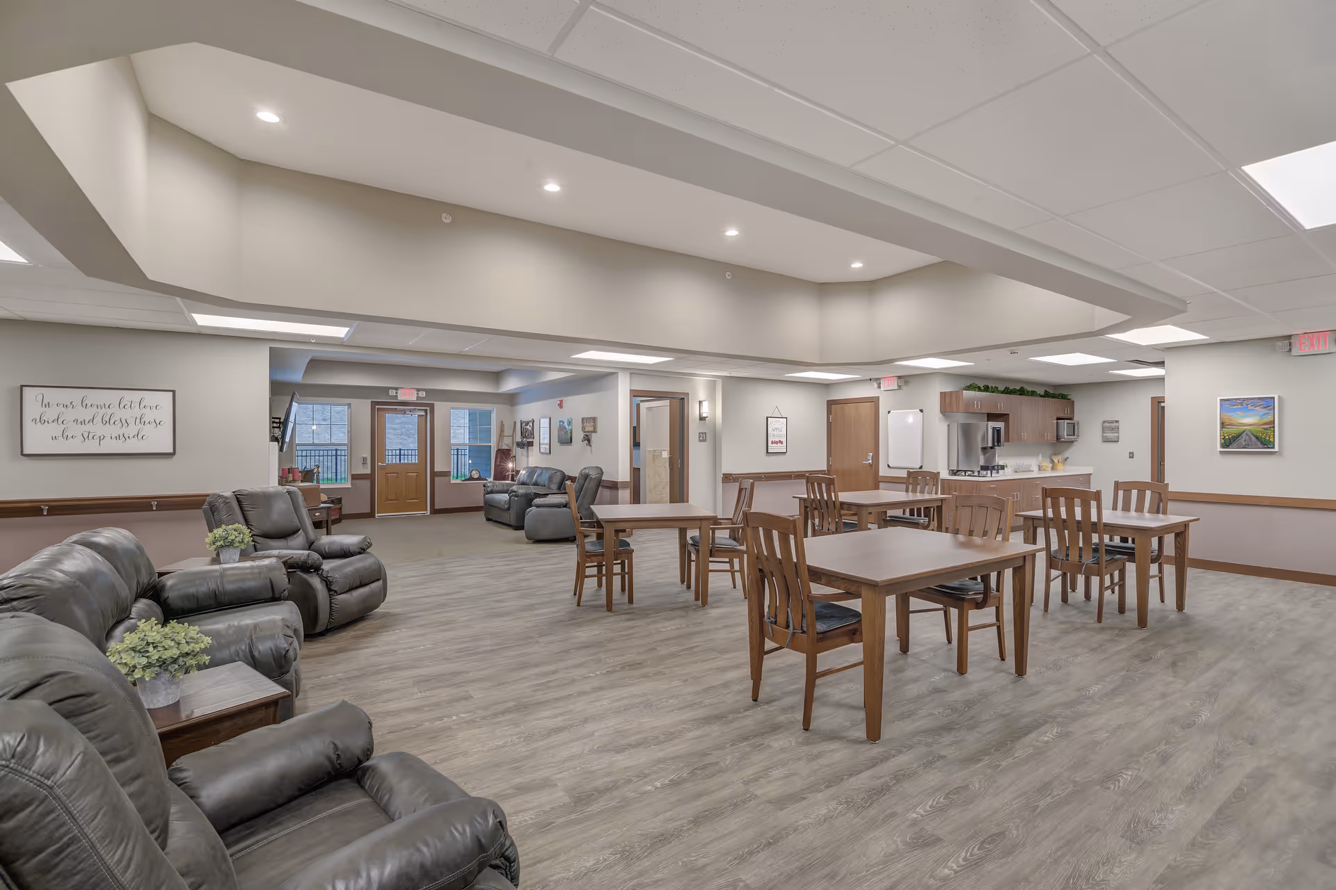 A spacious senior living common area featuring several wooden tables with chairs arranged for dining or activities. To the left, there are multiple black leather recliners with small tables and potted plants. The room has light-colored walls, wood trim, and a ceiling with recessed lighting. In the background, there is a small kitchenette area with cabinets, a refrigerator, and a microwave. The space is well-lit with natural light coming through windows and doors.