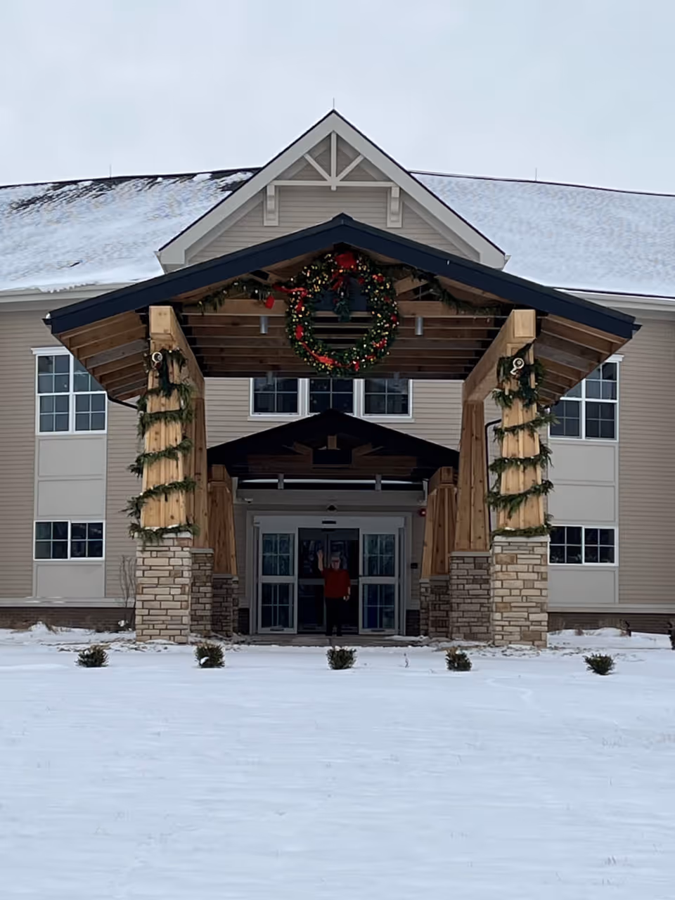 Snow-covered front entrance of a multi-story building with a wooden porte-cochere decorated with a large Christmas wreath and garlands.