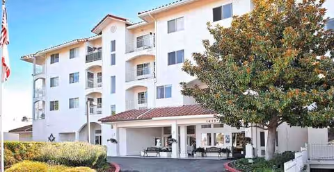 White multi-story senior living building with balconies, a canopy entrance drive, and mature trees.