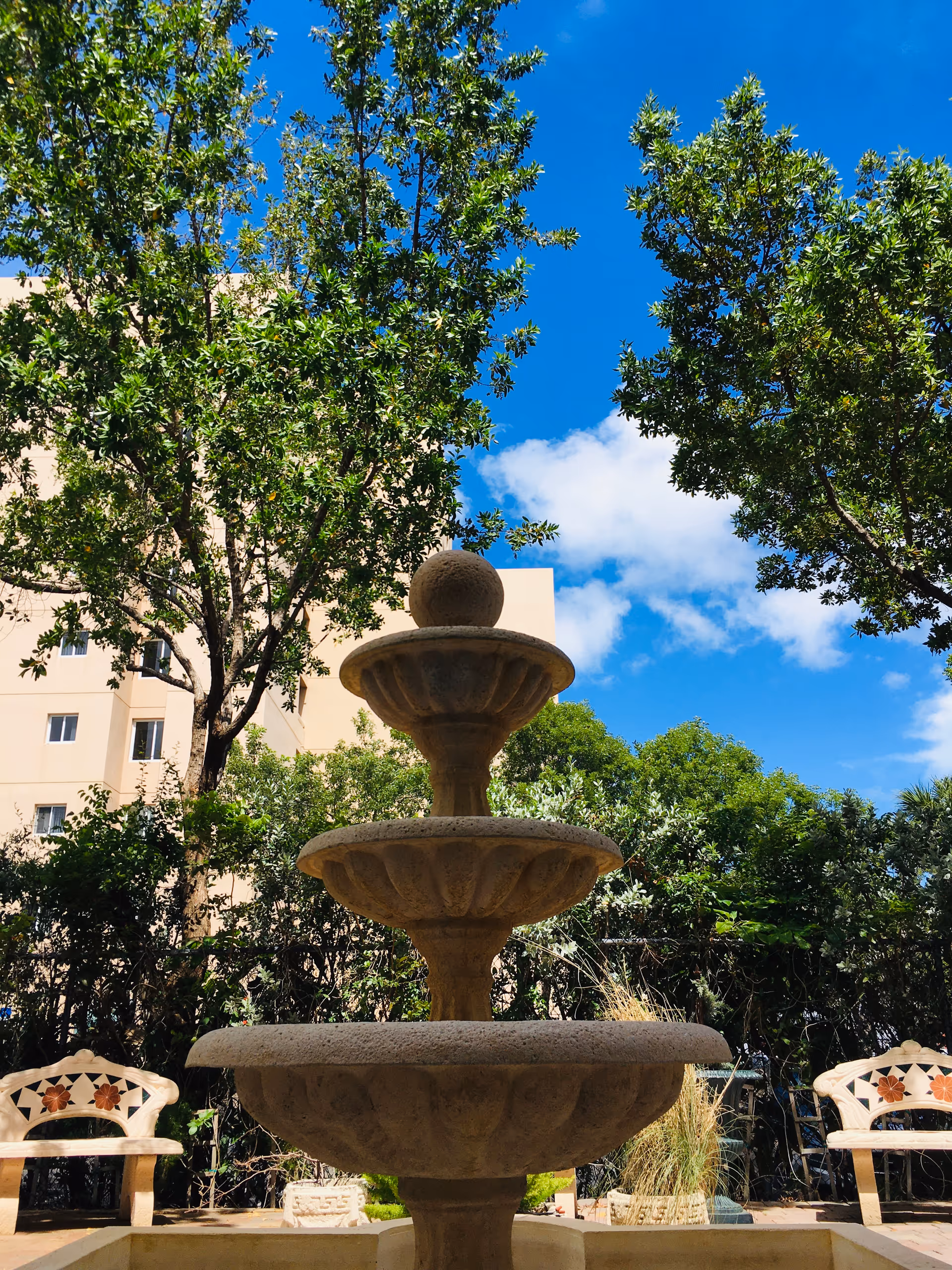Outdoor garden area with a three-tier stone fountain in the center, surrounded by green trees and bushes. Two decorative benches with floral designs are visible on either side of the fountain. A beige building is partially visible in the background under a bright blue sky with some white clouds.