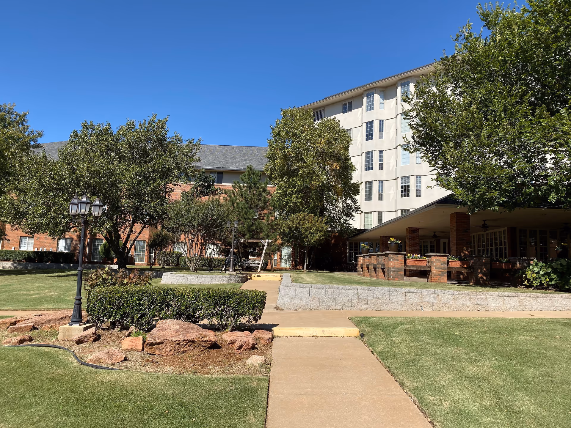 Outdoor view of Tealridge Retirement Community showing a paved walkway leading through a landscaped garden area with trimmed bushes, trees, and a lamp post. The background features a multi-story building with large windows and a covered patio area with brick pillars.