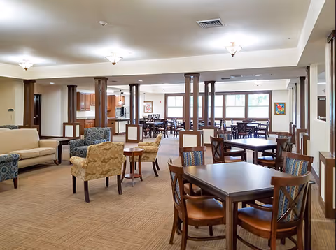A spacious senior living common area with multiple seating arrangements including armchairs, sofas, and wooden dining tables with chairs. The room features beige carpeting, wooden pillars, and large windows letting in natural light. The background shows a kitchen area and framed artwork on the walls.