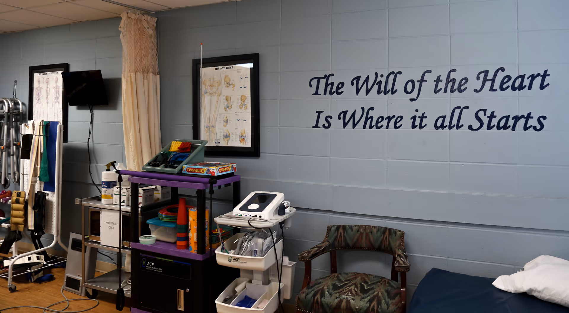 A rehabilitation therapy room with medical equipment, therapy tools, a chair, and a bed. The wall has a motivational quote that reads 'The Will of the Heart Is Where it all Starts.'