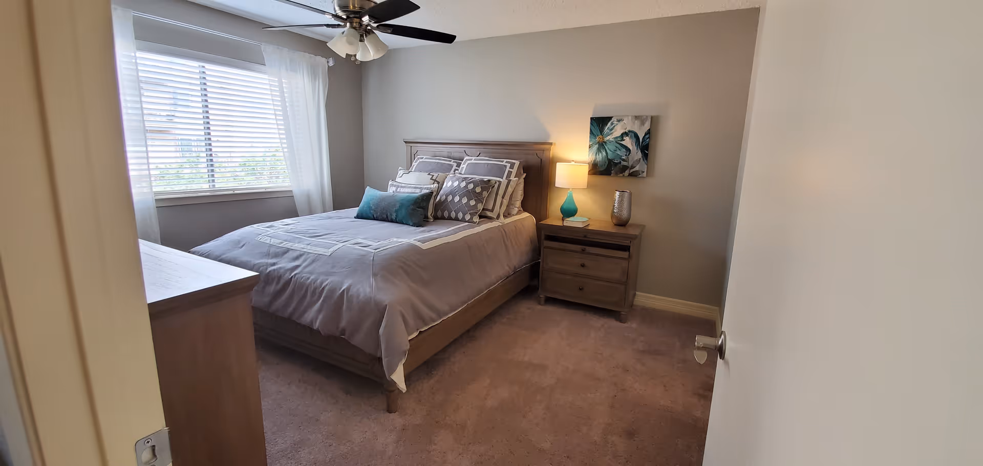 A cozy bedroom with a neatly made bed featuring multiple pillows and a gray bedspread. There is a wooden nightstand with a blue lamp, a book, and a decorative vase. A floral painting hangs on the wall above the nightstand. The room has beige walls, a ceiling fan with lights, a window with white blinds and sheer curtains, and a carpeted floor.