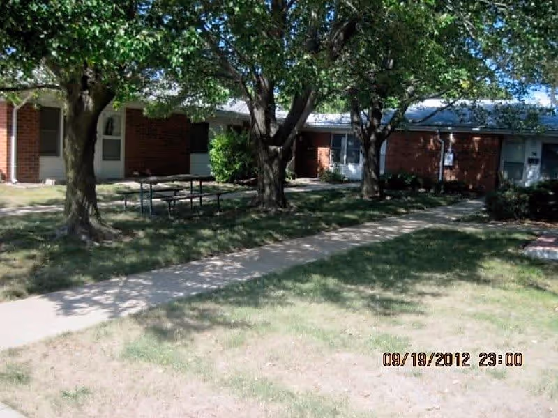 Outdoor area of Park View Senior Village featuring a grassy lawn with a concrete walkway, several mature trees providing shade, a picnic table, and a single-story brick building with multiple doors and windows in the background.
