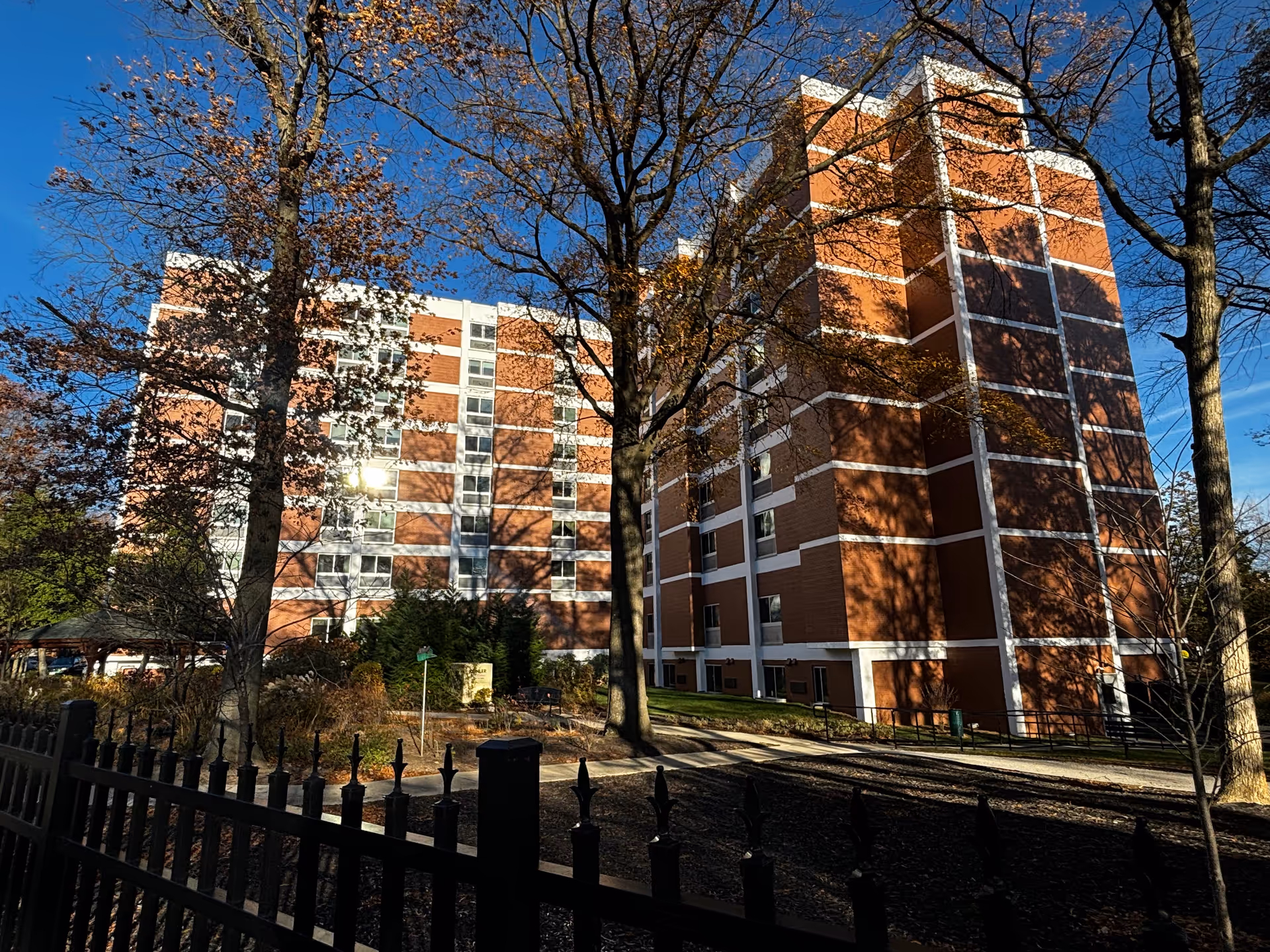Exterior view of a multi-story brick residential building with white trim, surrounded by leafless trees and a black metal fence in the foreground under a clear blue sky.