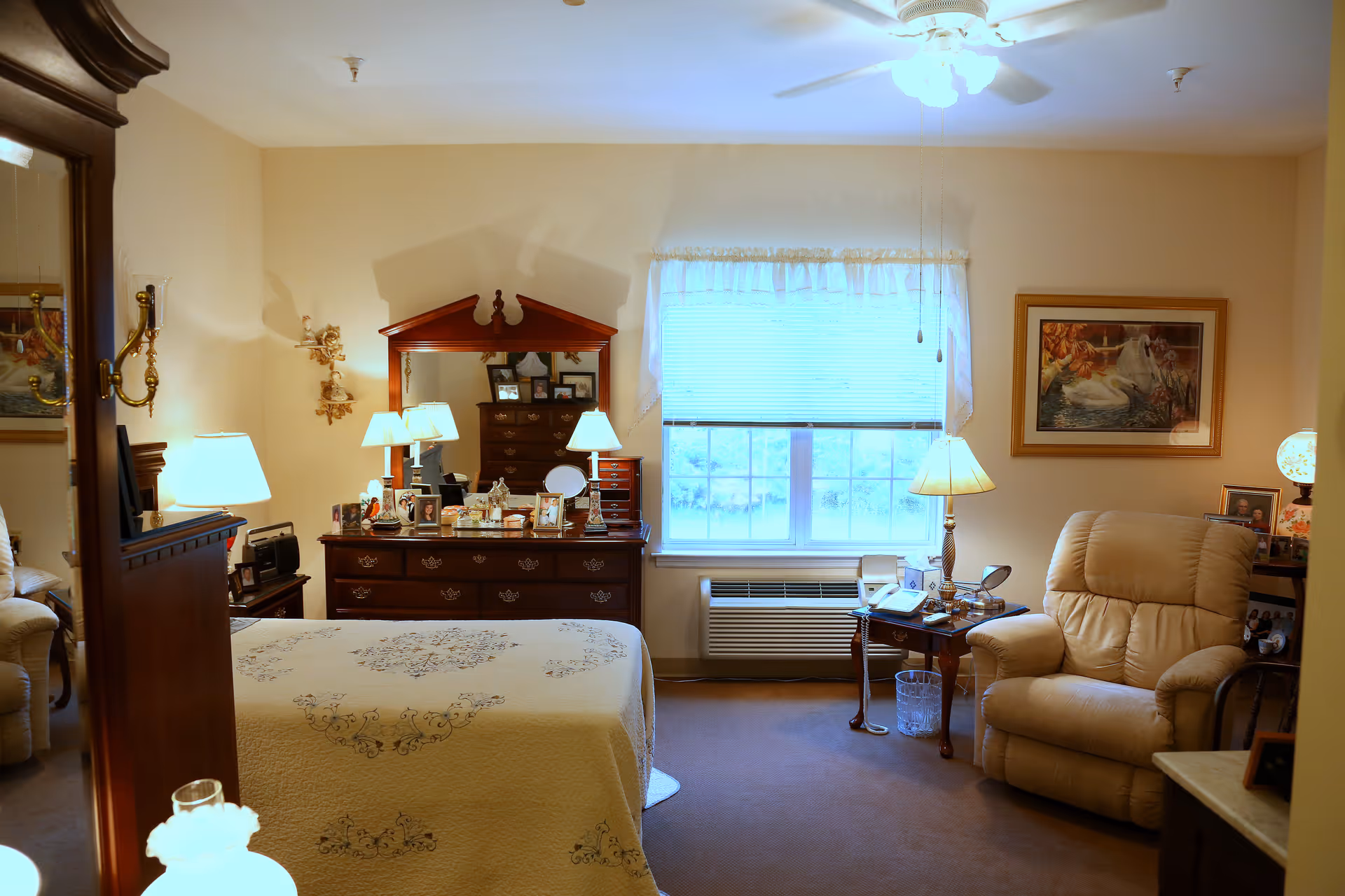 A cozy furnished bedroom with a bed in the foreground, a wooden dresser and mirror, an armchair, side tables and a window letting in daylight.