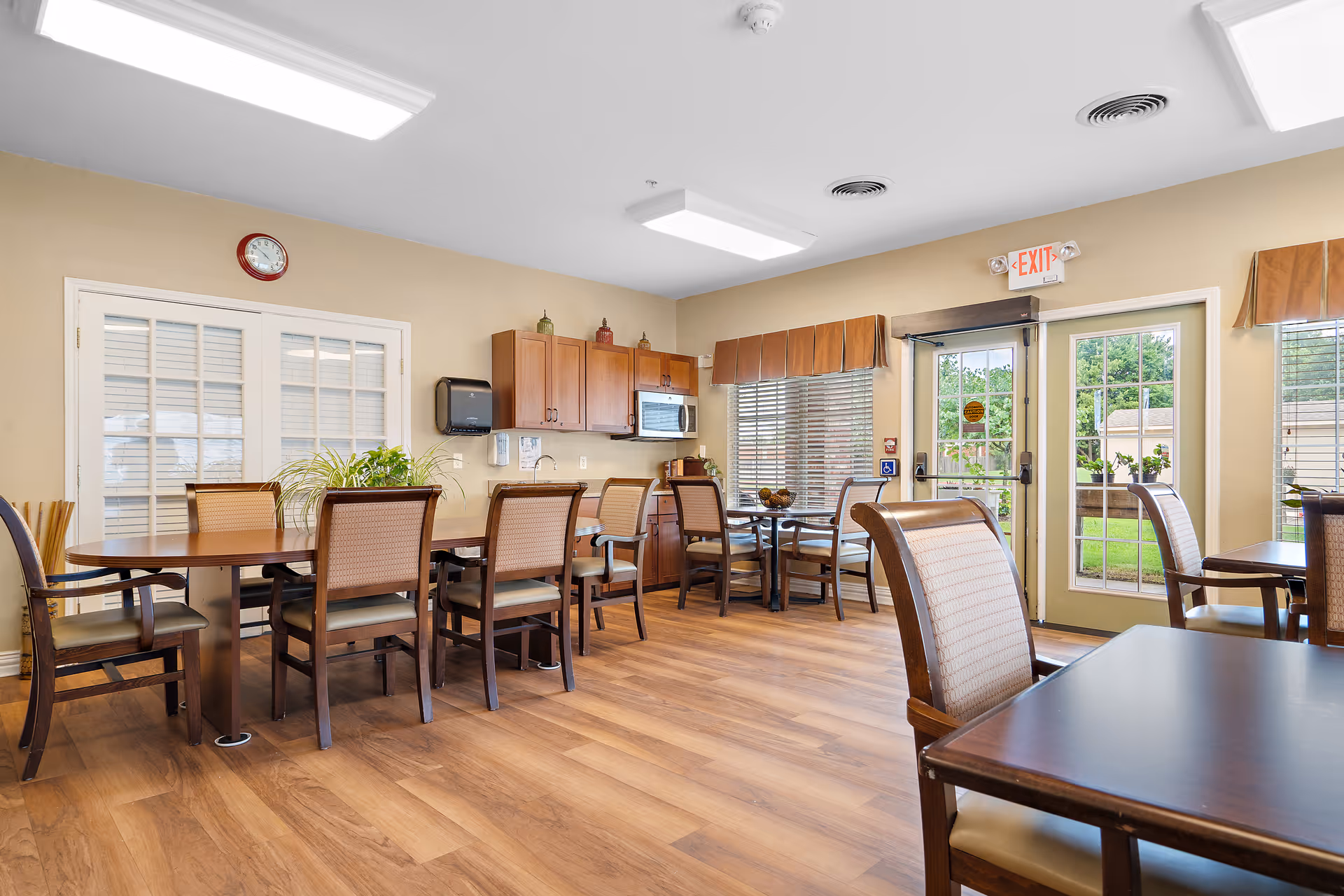 A bright dining room with wooden floors, multiple tables and chairs, a kitchenette with cabinets, microwave, and sink, large windows with blinds, and a glass door leading outside.