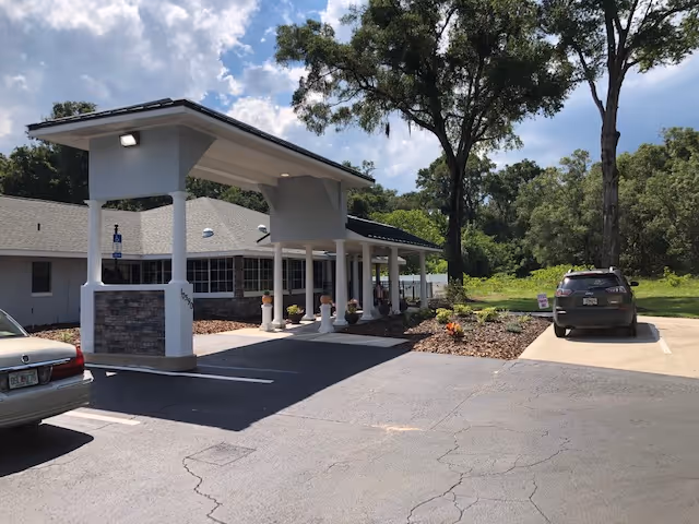Front entrance of a single-story assisted living building with a covered drive-up canopy, parked cars, and surrounding trees.