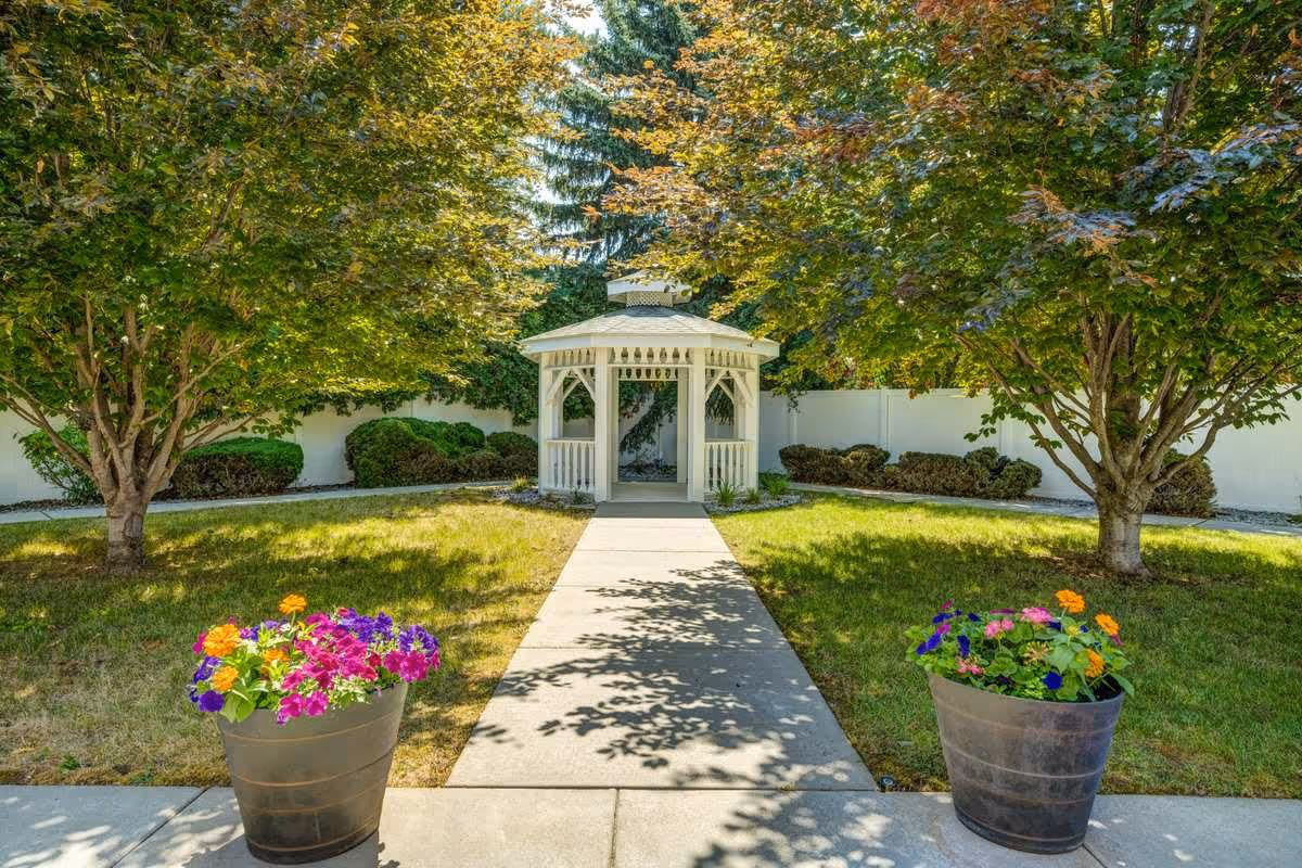 A peaceful garden area with a concrete walkway leading to a white gazebo surrounded by green grass, colorful flower pots, and trees with green and orange leaves.