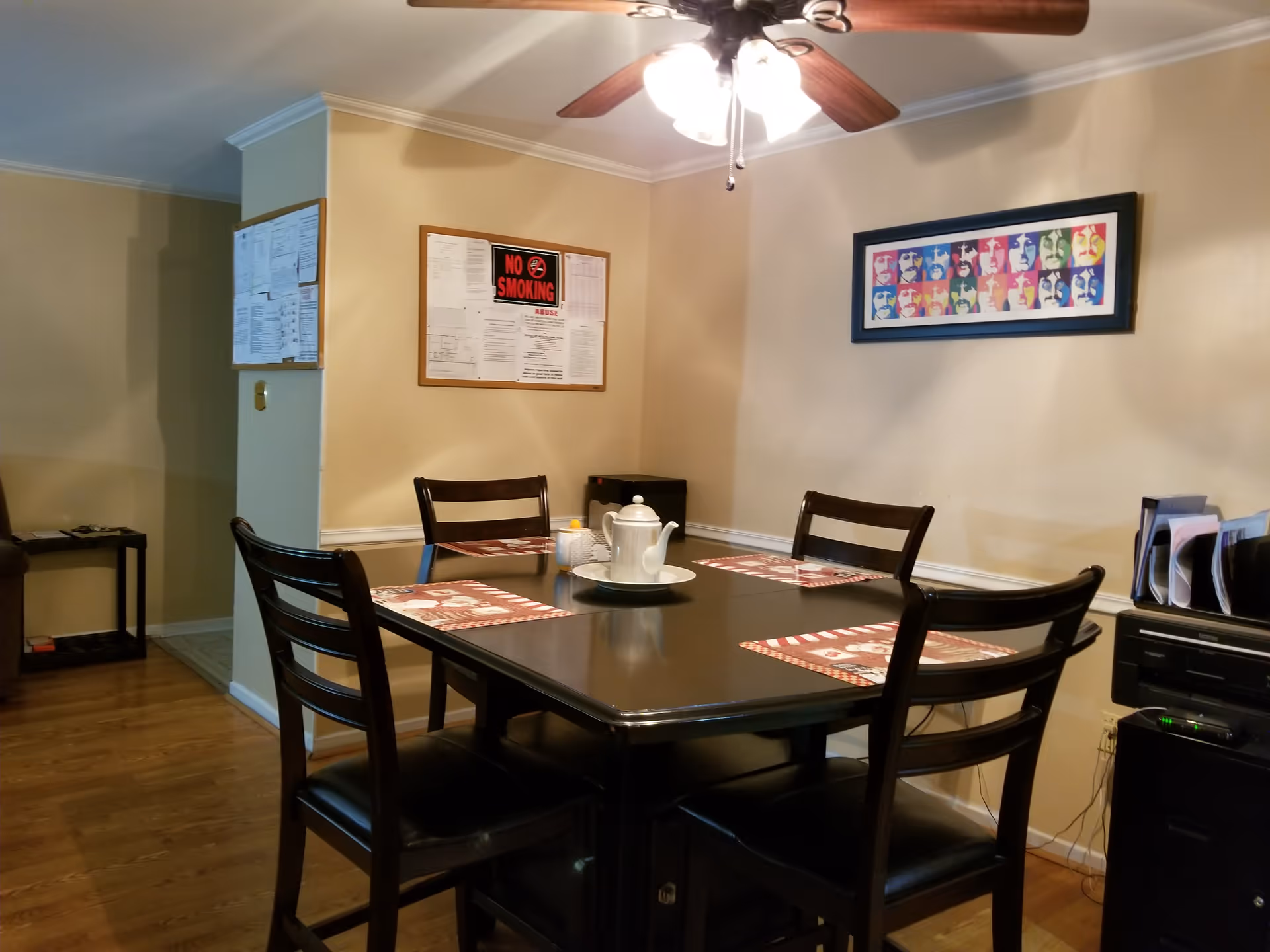 A dining area with a dark wooden table set for four with placemats and a white teapot on a tray in the center. Four matching chairs surround the table. The room has beige walls with a colorful framed artwork on one wall and a bulletin board with notices, including a 'No Smoking' sign, on another. A ceiling fan with lights is above the table. There is a small black cabinet and a printer with organized files on the right side of the room.