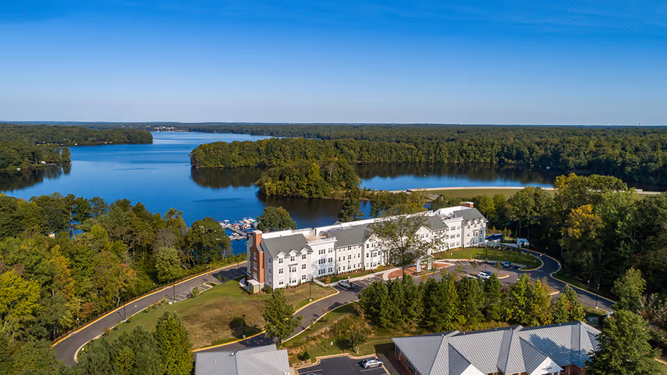 Aerial view of Auburn Hill Senior Living facility surrounded by trees and situated near a large lake with a dock and boats. The building is a multi-story structure with a parking area and curved driveway in front, set in a lush green landscape under a clear blue sky.
