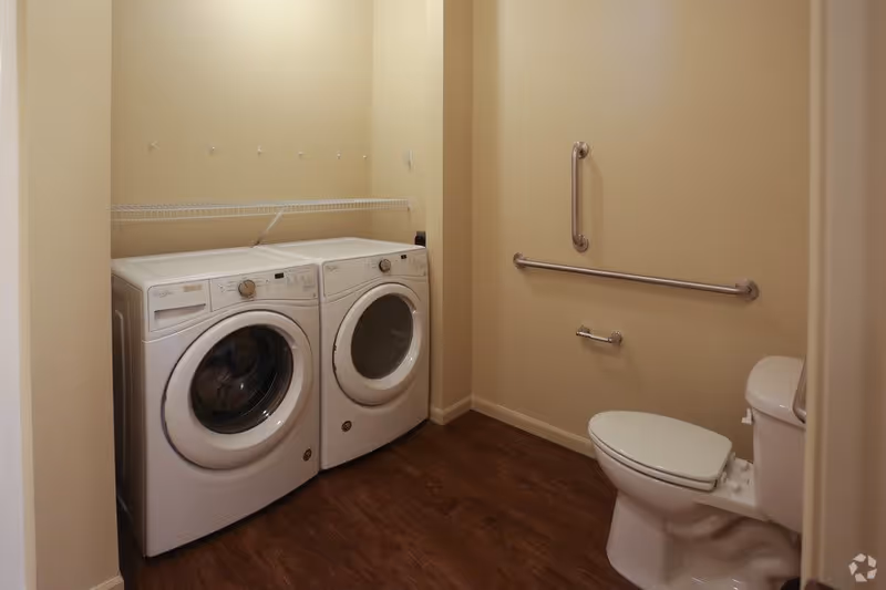 A small room with a white front-loading washing machine and dryer side by side on the left, beige walls, a white toilet on the right, and two metal grab bars mounted on the wall above and beside the toilet. There is a wire shelf above the washer and dryer and a wooden floor.