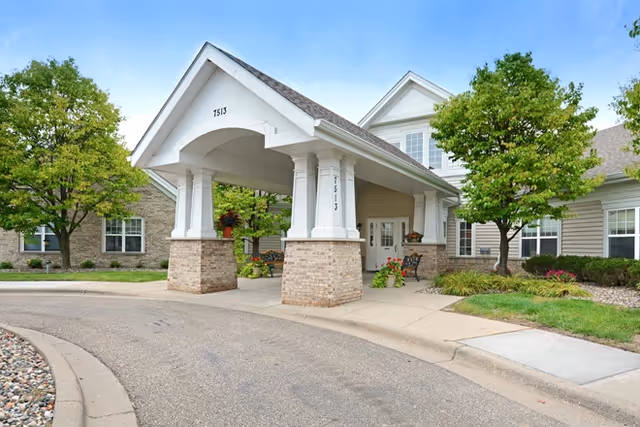 Entrance of a senior living facility with a covered driveway supported by brick and white columns, surrounded by green trees and landscaping under a clear blue sky.