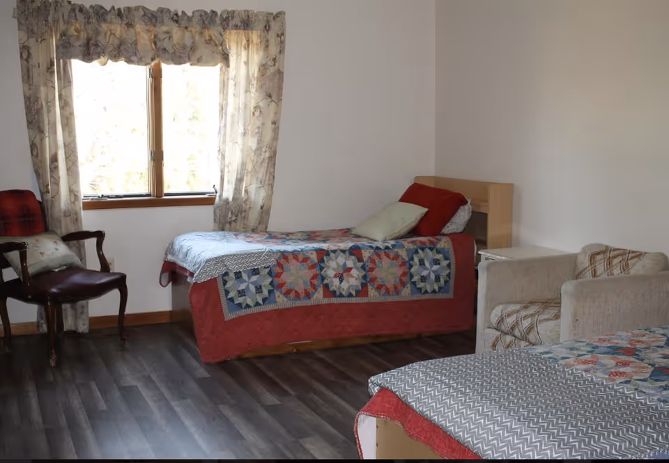 A simple bedroom with two twin beds covered in patterned quilts, a window with curtains, and a couple of chairs.