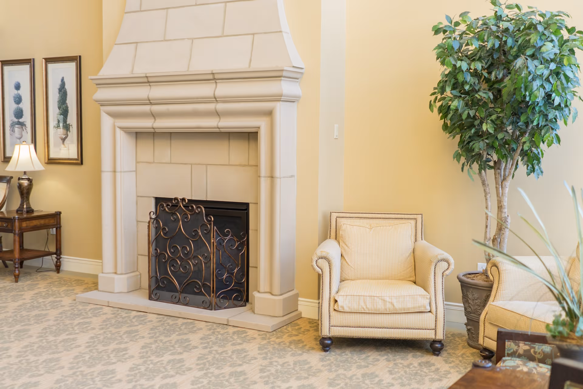 A cozy living room area featuring a beige armchair with a cushion, a large decorative fireplace with an ornate metal screen, a potted indoor tree, a wooden side table with a lamp, and framed botanical artwork on the wall. The room has light yellow walls and patterned carpet flooring.