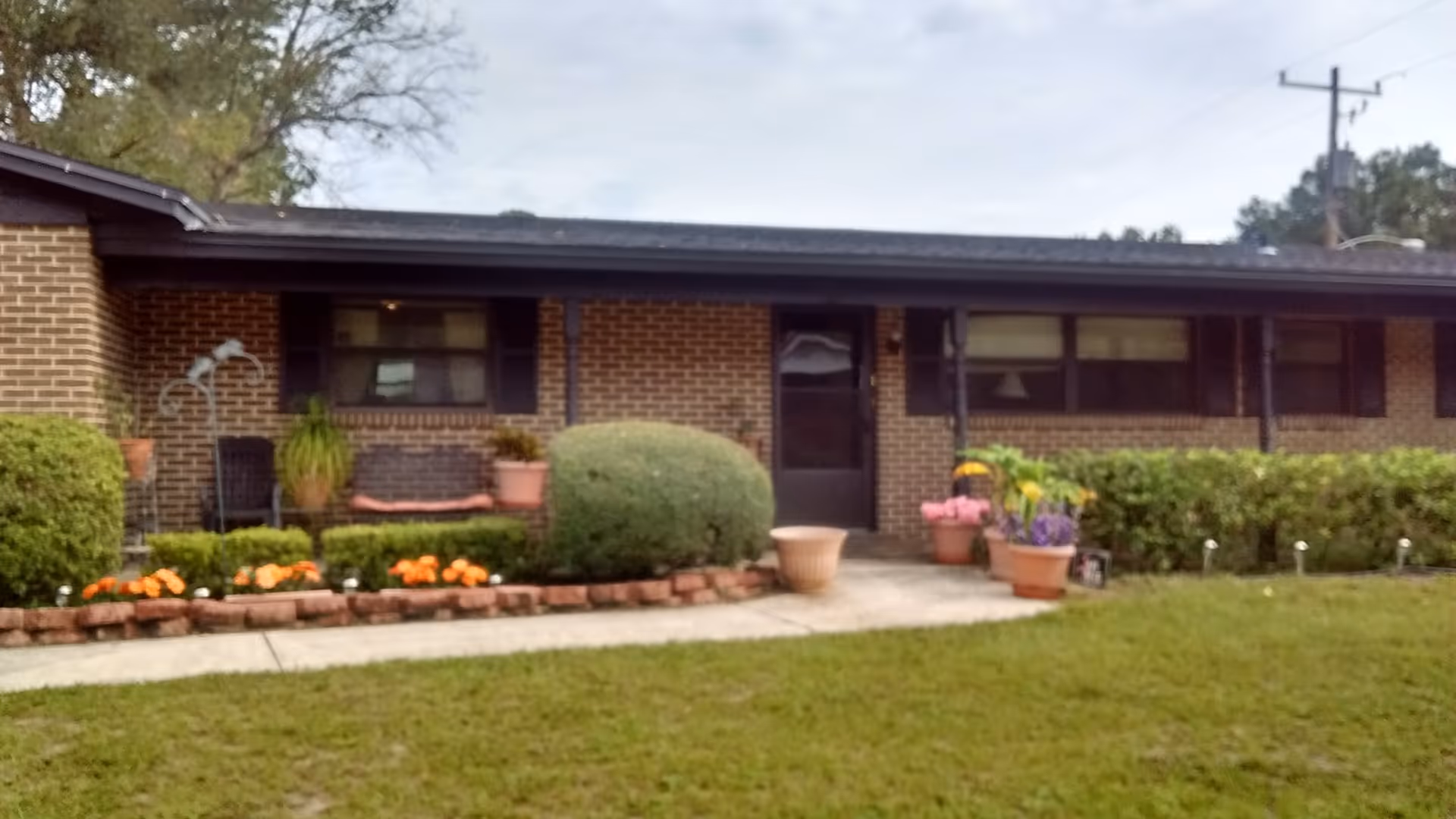 Single-story brick building with a black roof, a front door, and several windows. There are bushes, flower pots with colorful flowers, and a small garden bed with orange flowers along the front. A concrete walkway leads to the entrance. The sky is partly cloudy.