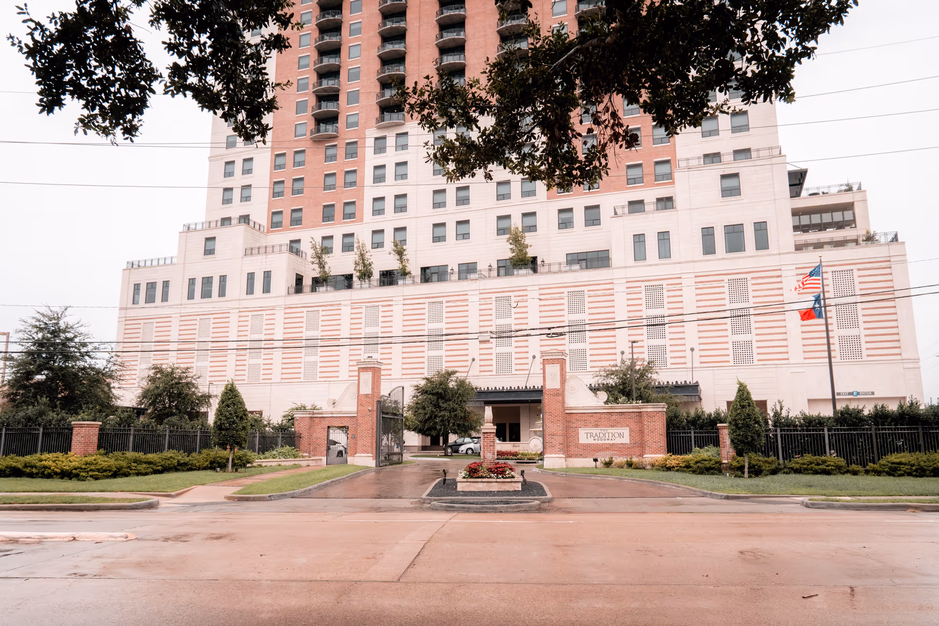 Front exterior view of The Tradition–Woodway building, a multi-story structure with a gated entrance, landscaped greenery, and American and Texas flags on flagpoles.