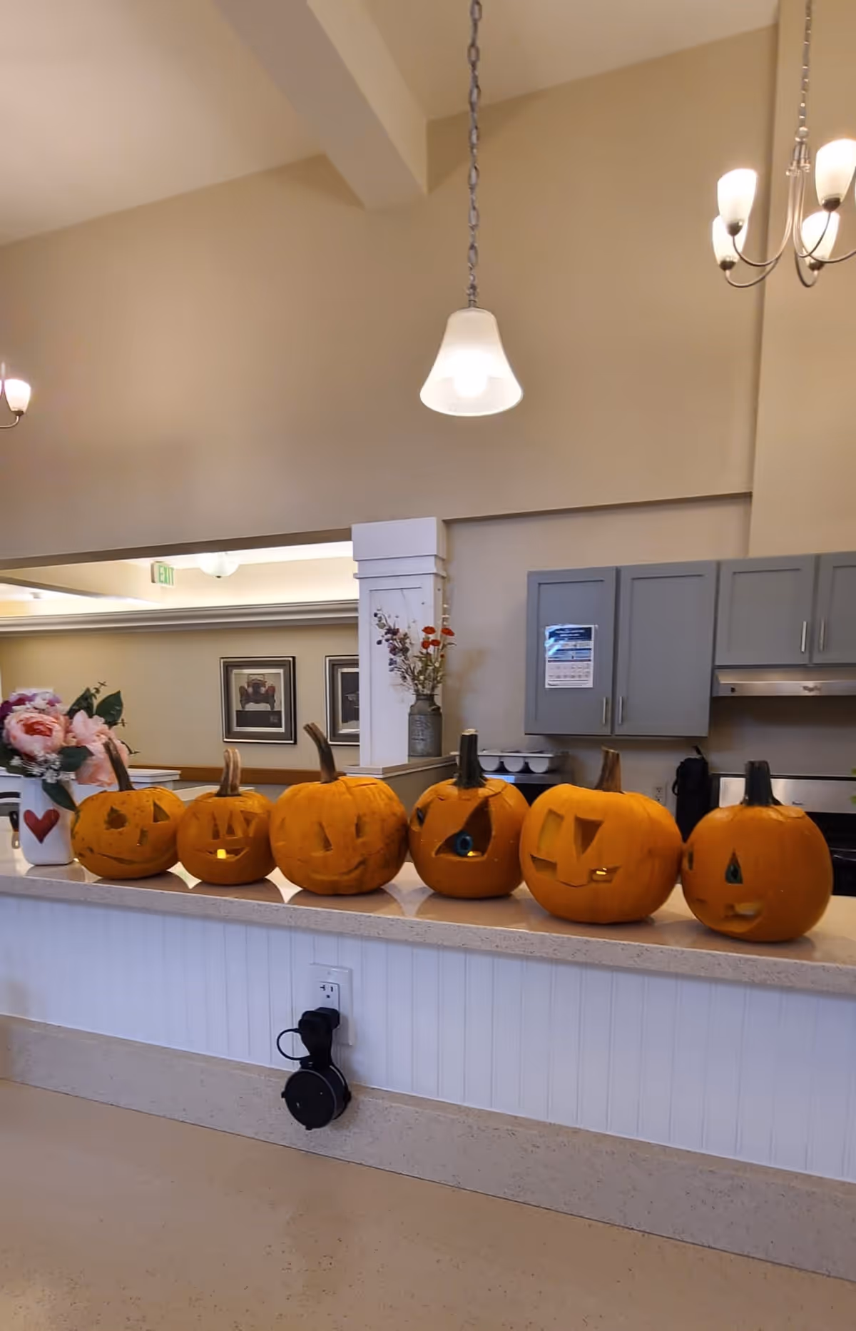 A row of six carved Halloween pumpkins with various facial expressions displayed on a countertop in a well-lit interior space. The background shows a kitchen area with gray cabinets, a stove, and a vase with flowers on a pillar. Pendant lights hang from the ceiling above.