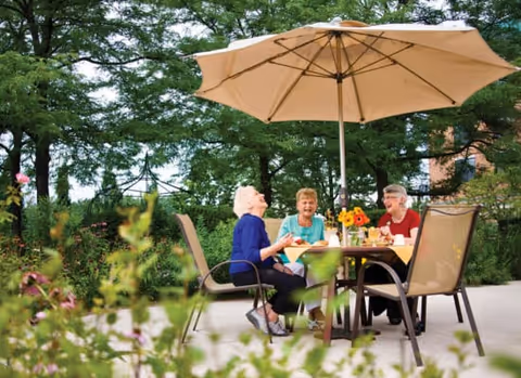Three elderly women sitting around a table outdoors under a large beige patio umbrella, enjoying conversation and drinks in a garden setting with green trees and plants in the background.