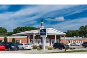 Exterior view of The Meadows on University senior living facility showing a single-story brick building with a white entrance canopy and a small steeple on the roof. Several cars are parked in front under a partly cloudy sky.