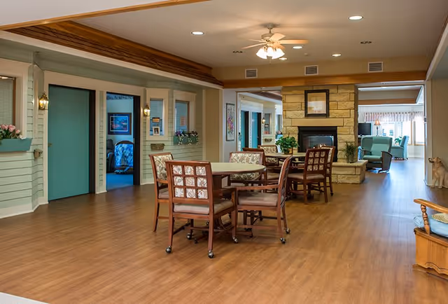 A spacious senior-living common area with round tables and chairs, a stone fireplace, and lounge seating in the background.