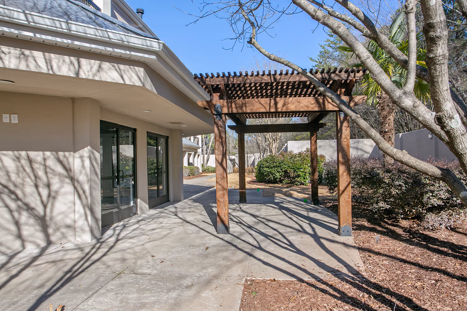 Outdoor view of a senior living facility showing a concrete walkway next to a beige building with glass doors. A wooden pergola structure is positioned on the walkway, surrounded by trees and shrubs under a clear blue sky.