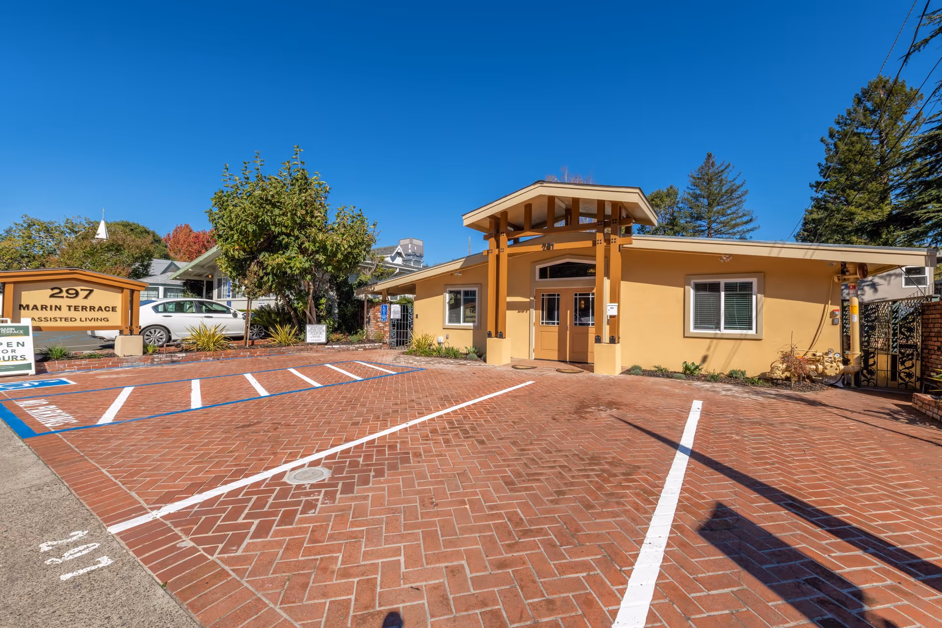 Front entrance and brick parking area of a single-story yellow assisted living building with a sign reading '297 Marin Terrace Assisted Living'.