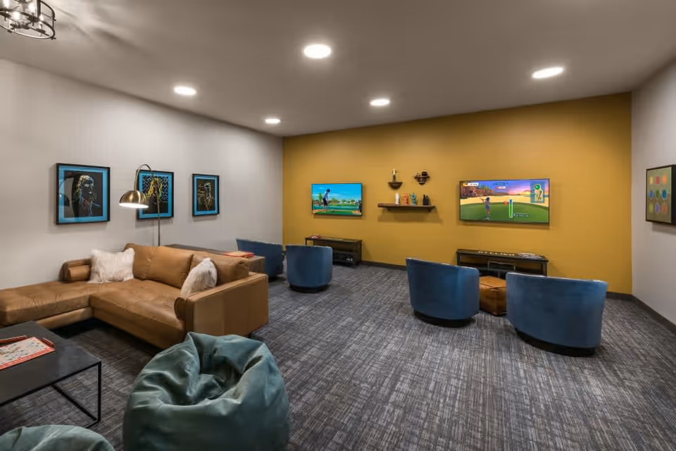 A modern lounge area with a brown leather sectional sofa, blue round chairs, and green bean bags. The room has a gray carpet and a mustard yellow accent wall with two flat-screen TVs mounted, displaying a golf video game. There are framed artworks on the walls and a floor lamp beside the sofa.