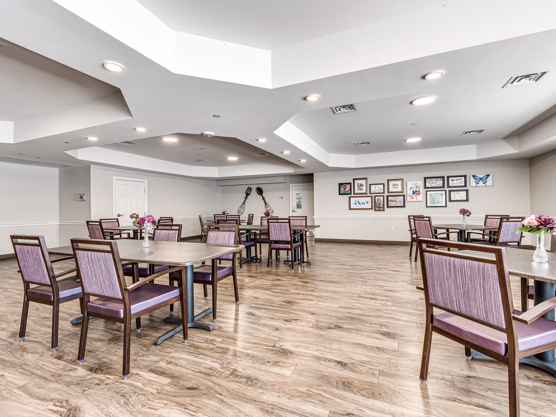 A spacious dining room with several square tables and purple cushioned chairs. Each table has a small vase with purple flowers. The room has light-colored walls, a wooden floor, recessed ceiling lights, and a wall decorated with framed pictures and large decorative utensils.