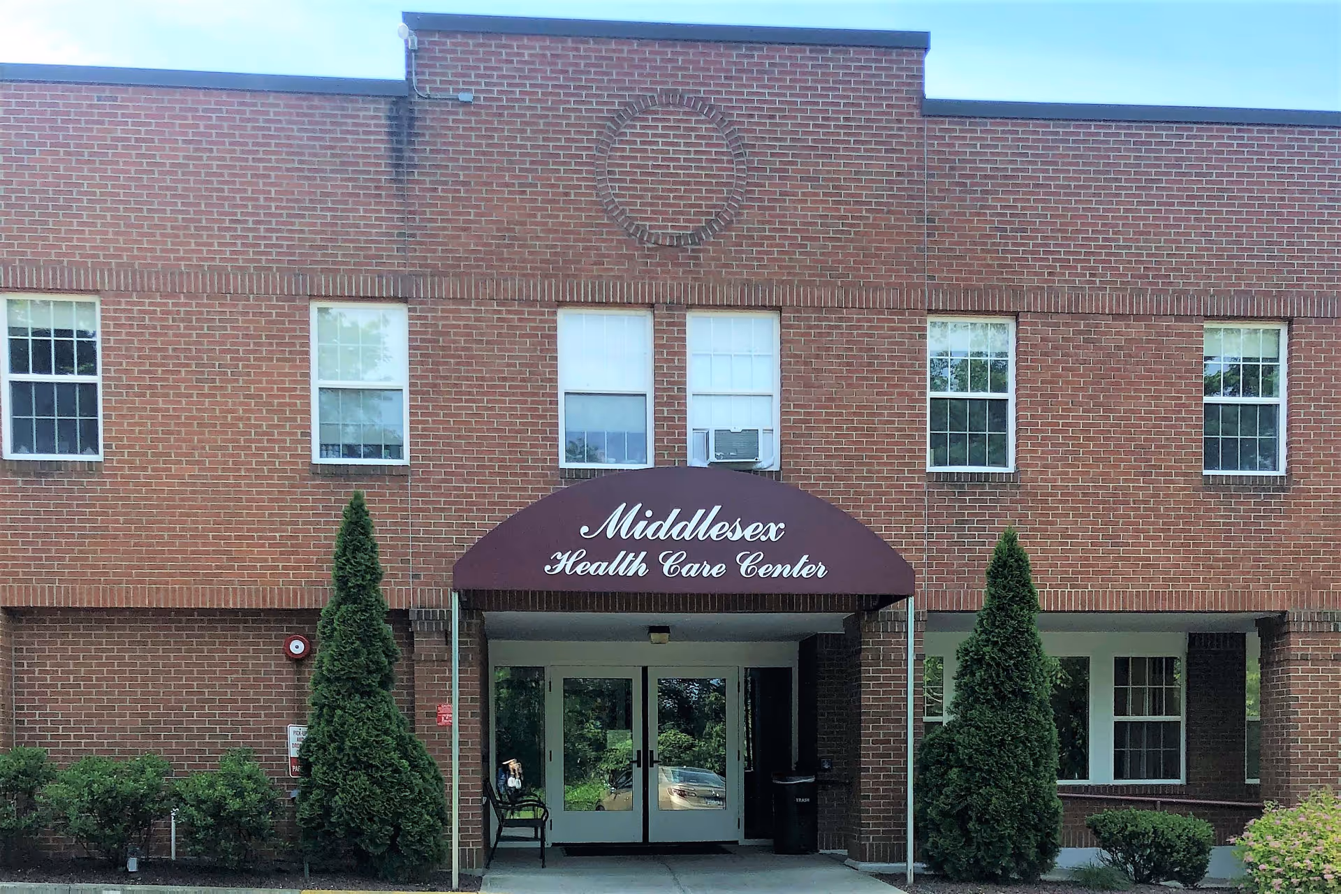 Front exterior view of a two-story brick building with a maroon awning that reads 'Middlesex Health Care Center'. There are several windows on the upper and lower floors, two tall evergreen shrubs flanking the entrance, and a bench near the doorway.