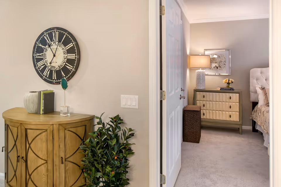 View of a hallway corner with a wooden cabinet topped with decorative items including books and a vase, a large round wall clock above it, and a green leafy plant beside it. Through an open door, a bedroom is visible with a dresser, a table lamp, a framed picture on the wall, and part of a bed with a tufted headboard.