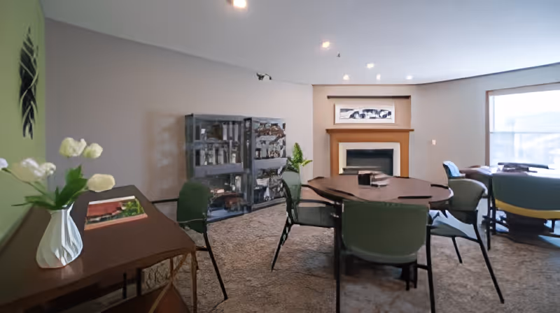 A cozy common room with a large wooden table surrounded by green chairs. There is a fireplace with a framed picture above it on the far wall. To the left, there is a wooden side table with a white vase holding white flowers. A glass-front cabinet with various items inside is against the back wall. Large windows on the right side let in natural light, and additional seating is visible near the windows.