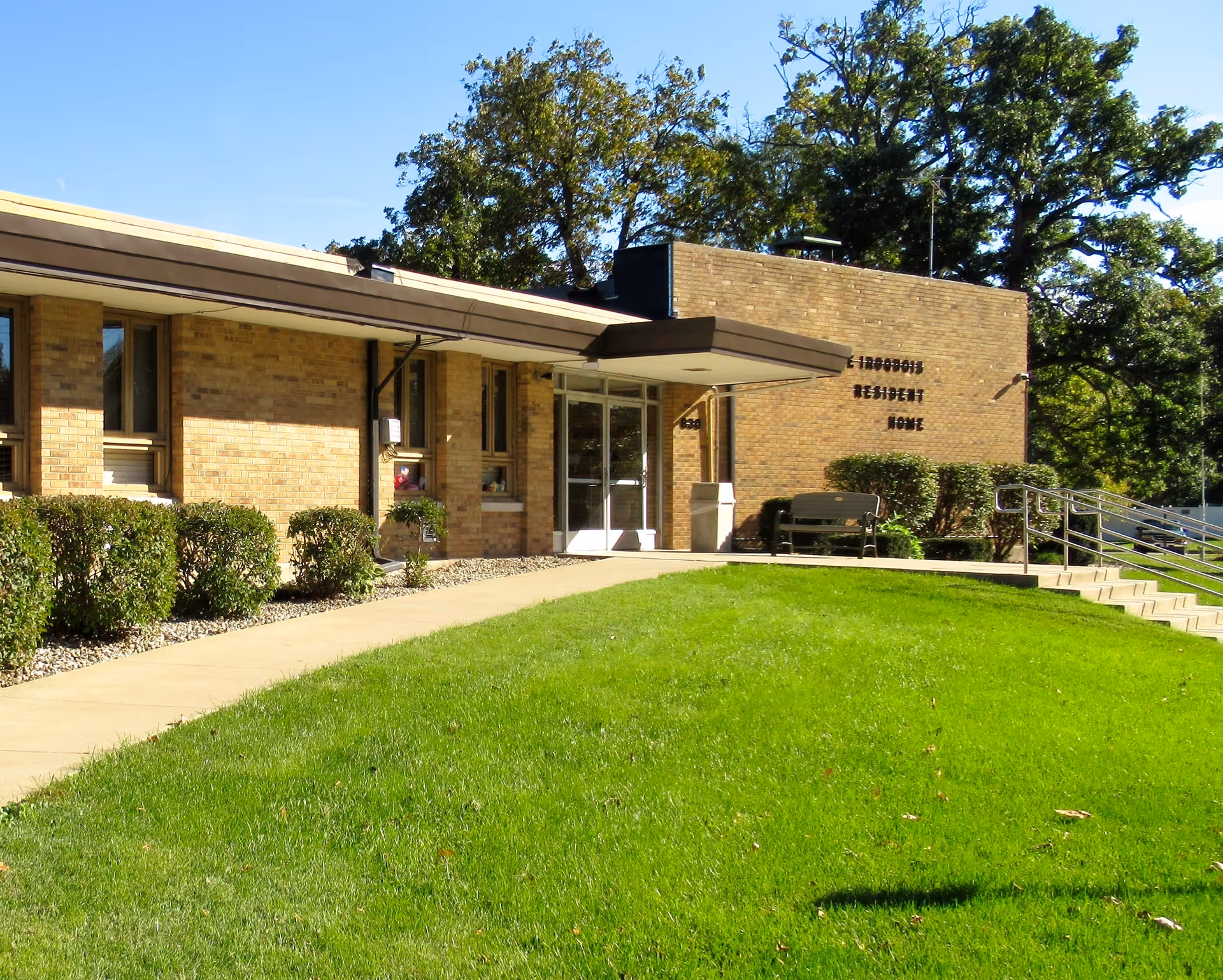 Exterior view of a single-story brick building with a sign that reads 'Iroquois Resident Home'. The building has several windows, a glass entrance door with a small overhang, a concrete walkway, green grass lawn, bushes along the building, and trees in the background under a clear blue sky.
