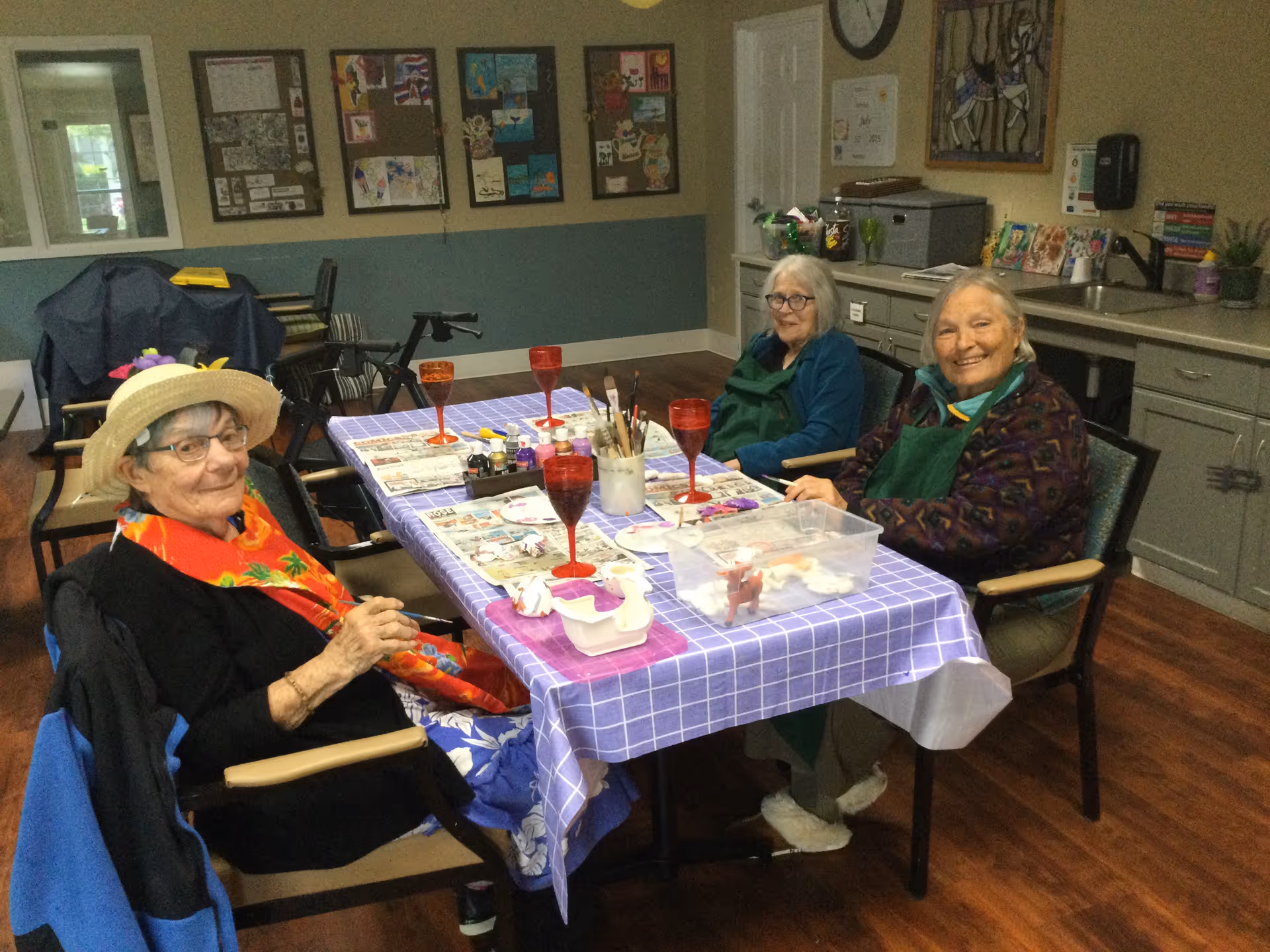 Three elderly women sitting around a table covered with a purple checkered tablecloth, engaged in an arts and crafts activity with paintbrushes, paints, and small figurines. The room has a cozy interior with cabinets, a sink, and artwork displayed on the walls.