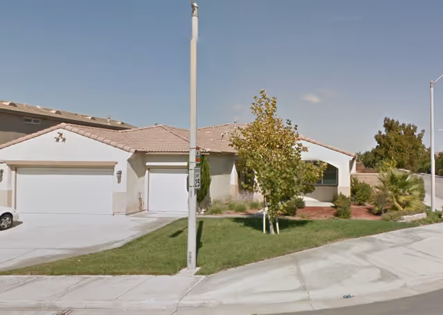 Single-story residential building with a tiled roof, a two-car garage, a driveway, and a small front yard with grass, trees, and shrubs under a clear blue sky.