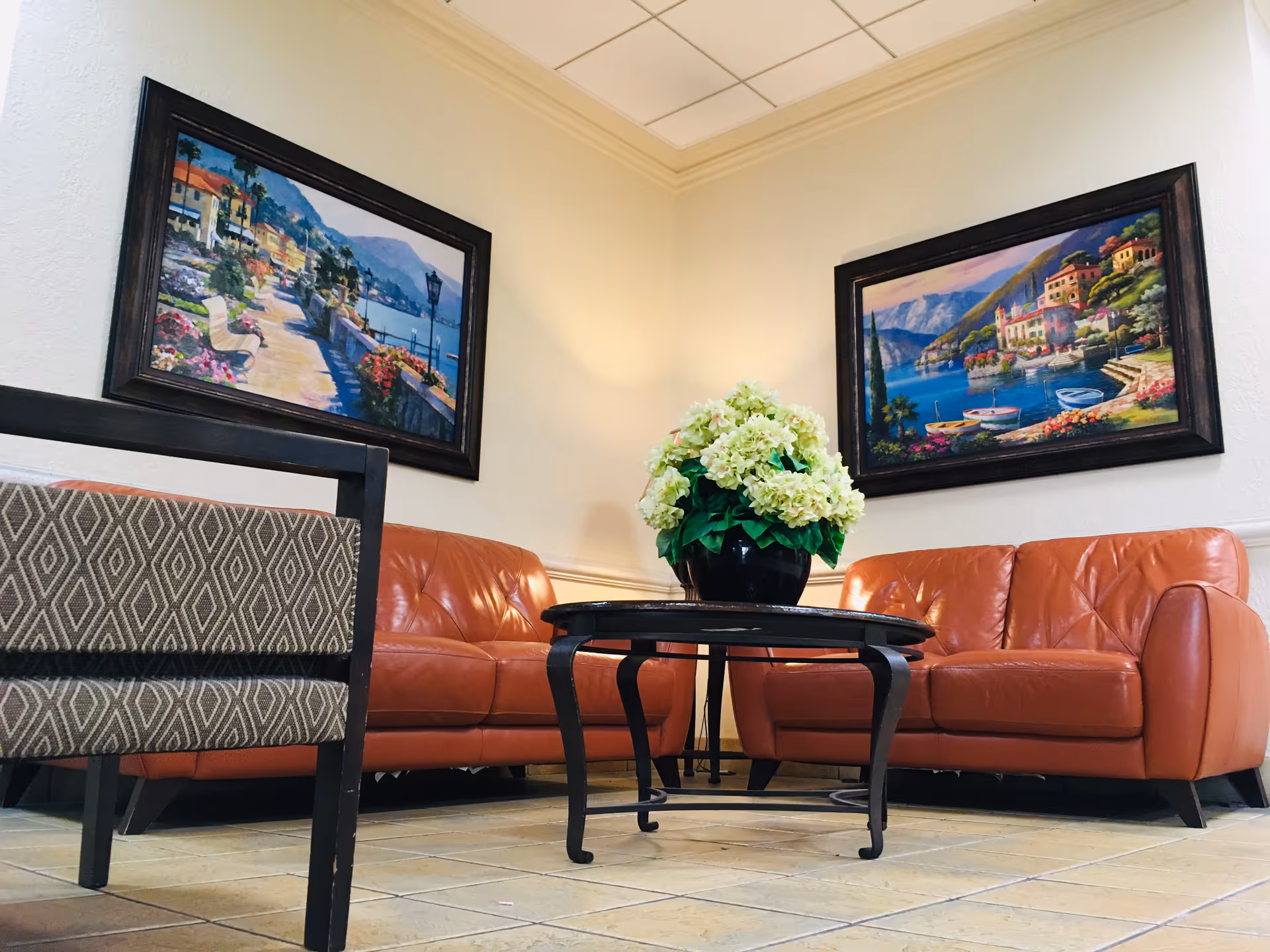 A cozy seating area with two orange leather sofas and a patterned armchair around a black coffee table with a potted plant with white flowers. Two framed paintings of scenic waterfront villages hang on the cream-colored walls.